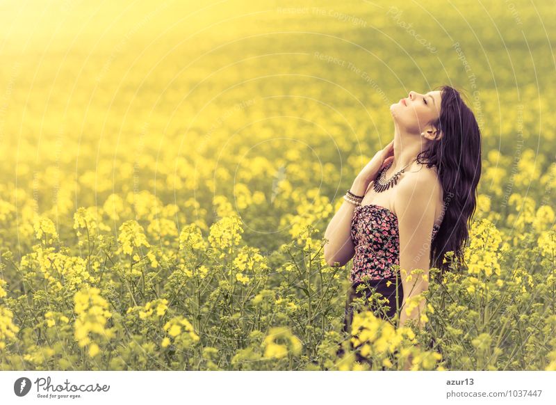 Schöne junge Frau genießt die warme Sonne in der Natur. Das attraktive Mädchen befindet sich in einer gelben grünen Wiese voller Blumen. Mit Kopf nach oben und geschlossenen Augen freut sie sich über den Frühling oder Sommer.