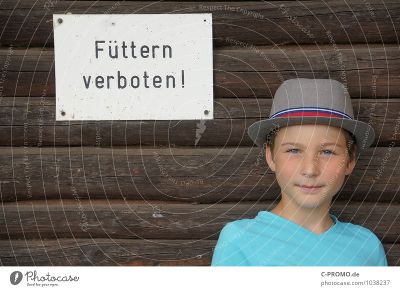Junge mit Hut steht an Holzwand mit Schild "Füttern verboten" Mensch maskulin Kind Kindheit 1 8-13 Jahre Hinweisschild Warnschild braun türkis selbstbewußt