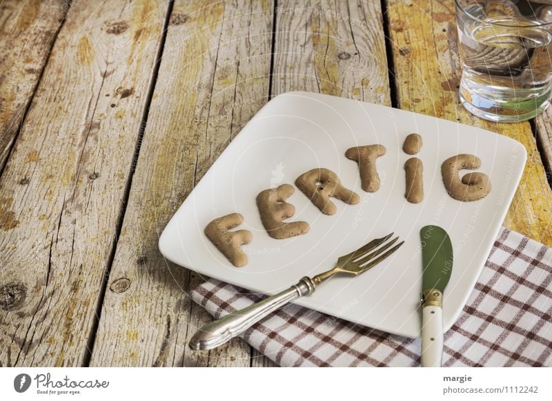 Die Buchstaben FERTIG auf einem Teller mit Messer und Gabel, Serviette, einem Glas Wasser auf einem rustikalen Holztisch Lebensmittel Frühstück Mittagessen