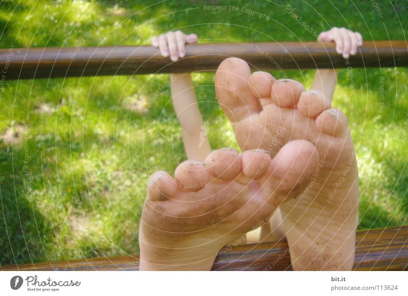 Kleines Kind hängt festgeklammert an einem Barren aus Holz, draussen in der Natur. Mädchen turnt an einer Stange, hält sich mit den Händen fest und streckt die nackten Füße nach oben. Lustige Füße, wirken durch Perspektive überdimensional groß und skurril.