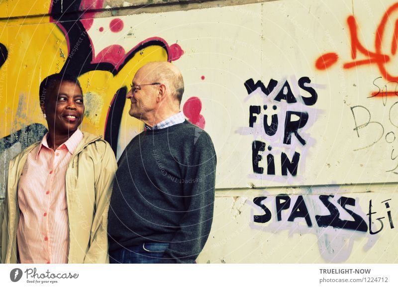 Schöne farbige Afrikanerin und weißer Senior bilden ein Paar mit Blick-Kontakt, das sich dicht nebeneinander stehend vor einer mit Graffiti und dem Text WAS FÜR EIN SPASS bemalten Wand liebevoll lächelnd in die Augen schaut