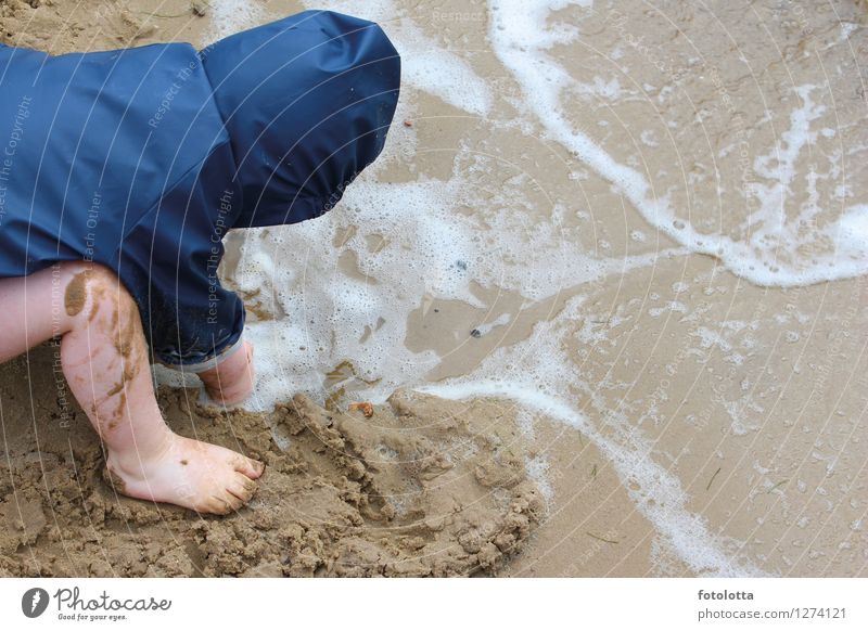 Buddeln Freude Spielen matschen Strand Kleinkind Beine Fuß Natur Sand Wasser Wellen Flussufer Regenjacke nass blau beige Matschepampe Barfuß Wind Außenaufnahme