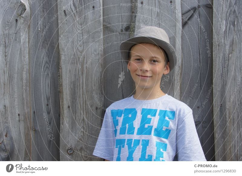 Junge mit Hut vor Holzzaun Mensch maskulin Kind 1 3-8 Jahre Kindheit 8-13 Jahre Mauer Wand T-Shirt Lächeln lachen frei Fröhlichkeit grau Coolness