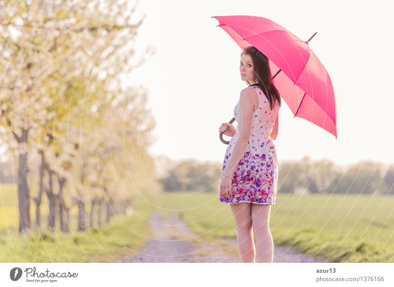 Frau mit Schirm bei wechselhaftem Wetter im Frühling oder Sommer. Die Wettervorhersage ist wechselhaft zwischen Sonne und Regen. Die attraktive junge Frau geht einen Spaziergang auf einem Weg in der Natur und wirft einen Blick zurück über die Schulter..