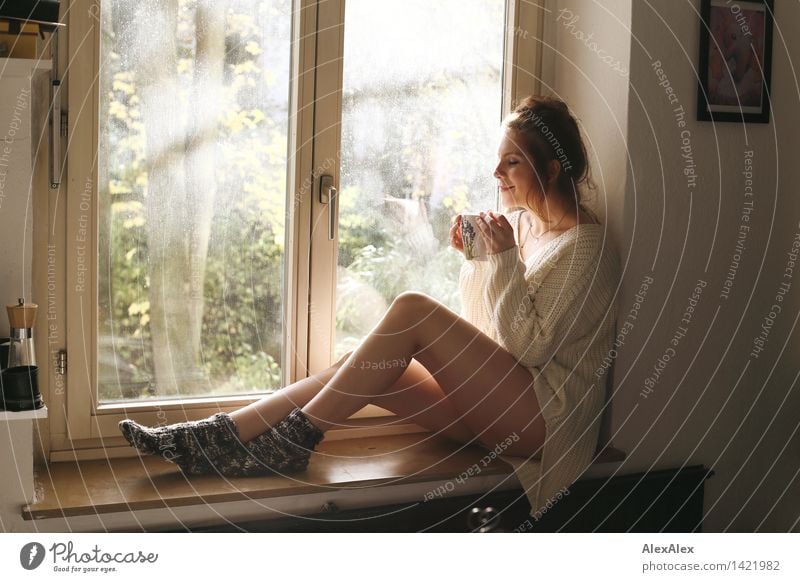Junge Frau sitzt in sonniger Küche im Fensterrahmen und genießt Kaffee oder Tee aus einer großen TasseSonntagskaffee trinken Becher harmonisch Erholung