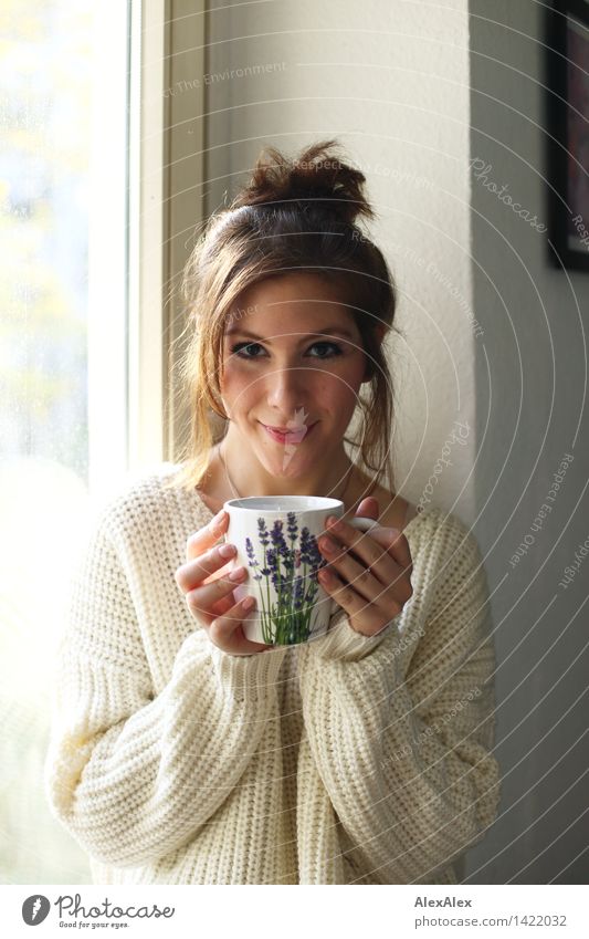 Junge Frau hält vor Fensterrahmen in der Küche eine Tasse in beiden Händen, die Sonntagskaffee enthält, sie lächelt in die Kamera Kaffee Frühstück trinken