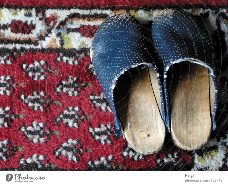 ein Paar abgenutzte Holzlatschen stehen auf einem alten gemusterten Teppich Schuhe Leder kaputt schäbig verschlissen Muster rot blau weiß Bekleidung obskur