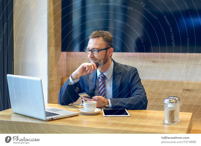 Geschäftsmann, der einen Kaffee genießt und mit einem Lächeln sein Handy auf Nachrichten überprüft, während sein Laptop und sein Tablet offen auf dem Tisch liegen