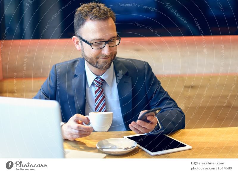 Geschäftsmann, der einen Kaffee genießt und mit einem Lächeln sein Handy auf Nachrichten überprüft, während sein Laptop und sein Tablet offen auf dem Tisch liegen