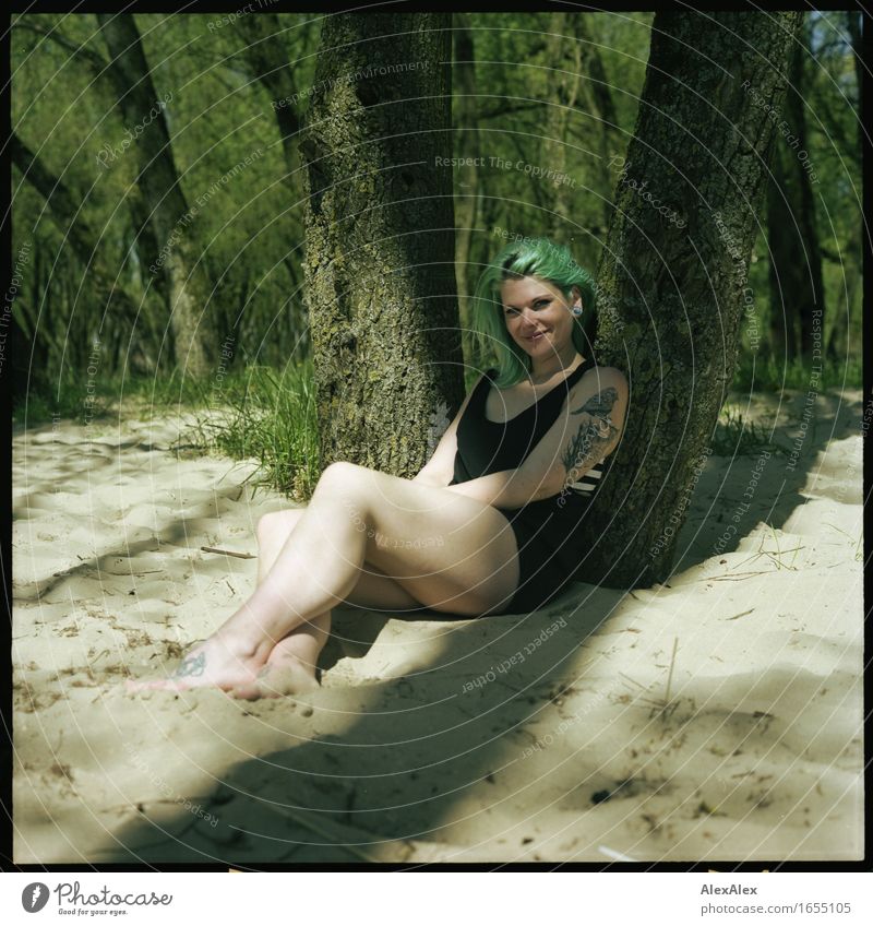Junge, tätowierte Frau mit türkisen Haaren sitzt barfuß im Strand- Wald an einen Baum gelehnt und lächelt in die Kamera Freude Erholung Sommerurlaub Sonnenbad