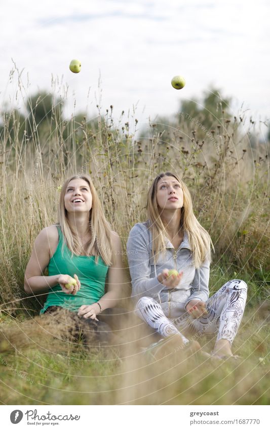 Zwei verspielte Mädchen mit langen blonden Haaren jonglieren im Freien, während sie Seite an Seite im langen Gras auf dem Land sitzen Frucht Apfel Glück schön