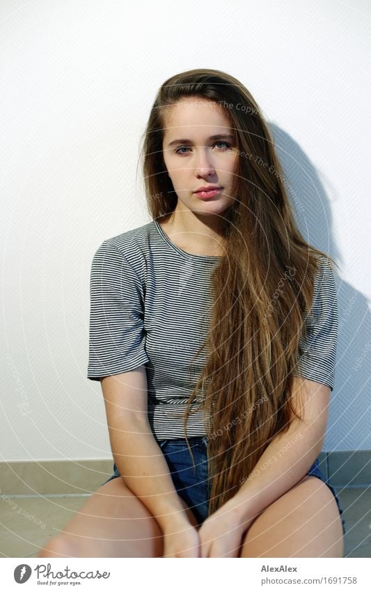 Junge, schöne Frau mit langen, dunkelblonden Haaren sitzt an einer weißen Wand und schaut in die Kamera Stil Junge Frau Jugendliche Haare & Frisuren 18-30 Jahre