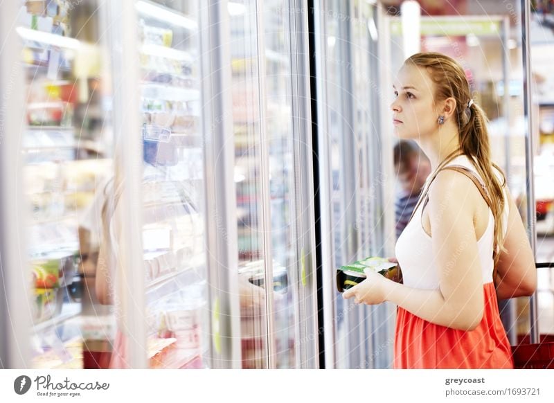 Junge Frau beim Kauf von Milchprodukten oder gekühlten Lebensmitteln im Supermarkt in der Kühlabteilung beim Öffnen der Glastür des Kühlschranks Lifestyle