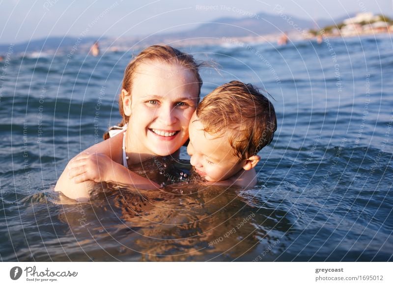 Glückliche attraktive junge Mutter mit einem schönen Lächeln, die ihrem kleinen Sohn das Schwimmen beibringt, während sie zusammen im Meer schwimmen Freude
