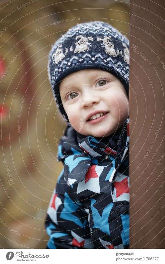 Niedlicher kleiner Junge in einer warmen Jacke und gestrickter Wintermütze, der mit einem freundlichen Lächeln um eine Tür herum in die Kamera blickt schön Kind