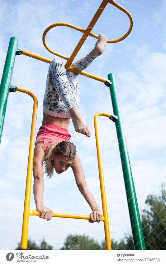 Schöne junge Turnerin arbeitet im Freien auf einem Satz von bunten Metallstangen hängen durch ihre Beine gegen einen bewölkten blauen Himmel Spielen Sport Kind