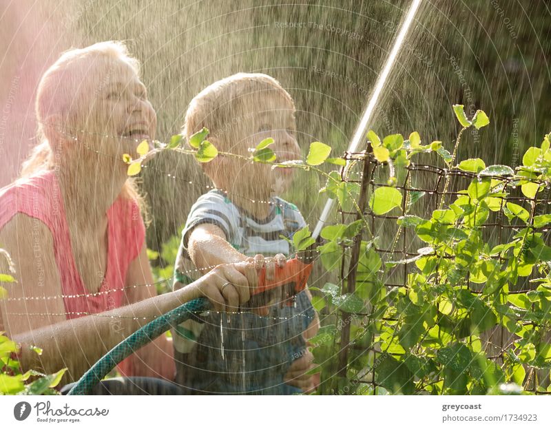 Lachende junge Mutter und ihr kleiner Sohn spielen mit einem Wasserstrahl aus einer Sprinklerdüse und lassen den Strahl über sich selbst in einem Sommergarten zurückfallen