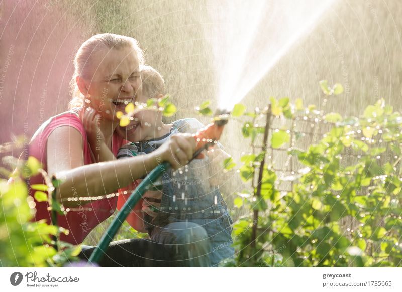 Lachende Mutter und Sohn, die mit einem Sprinkler im Garten spielen und den Strahl in die Luft lenken, so dass er zurückfällt und sie benetzt Freude Glück