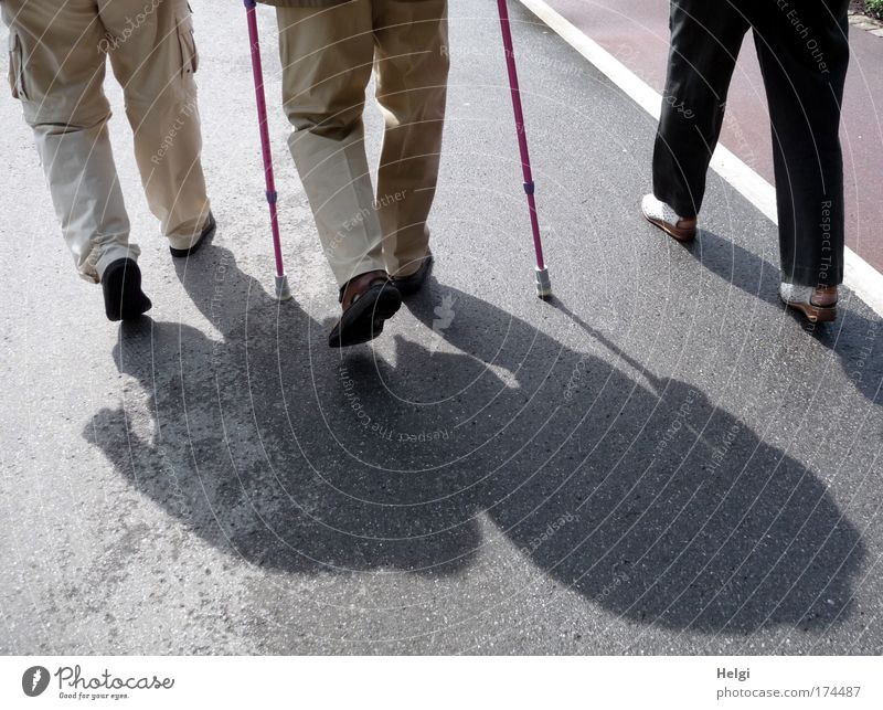 Beine von gehenden Senioren mit Stützen im Sonnenlicht mit Schatten auf einer Straße Farbfoto Gedeckte Farben Außenaufnahme Detailaufnahme Tag Licht Kontrast