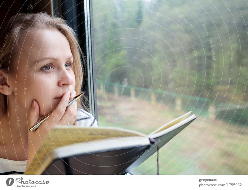 Junge Frau im Zug, die Notizen in ein Tagebuch oder Journal schreibt und nachdenklich aus dem Fenster starrt, während sie mit dem Stift an den Lippen überlegt, was sie schreiben soll.