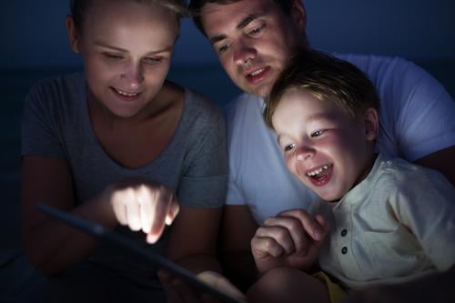 Glückliche dreiköpfige Familie mit Pad spät am Abend. Lachendes Kind schaut auf den Bildschirm, wo die Mutter auf etwas zeigt Freude Freizeit & Hobby Spielen
