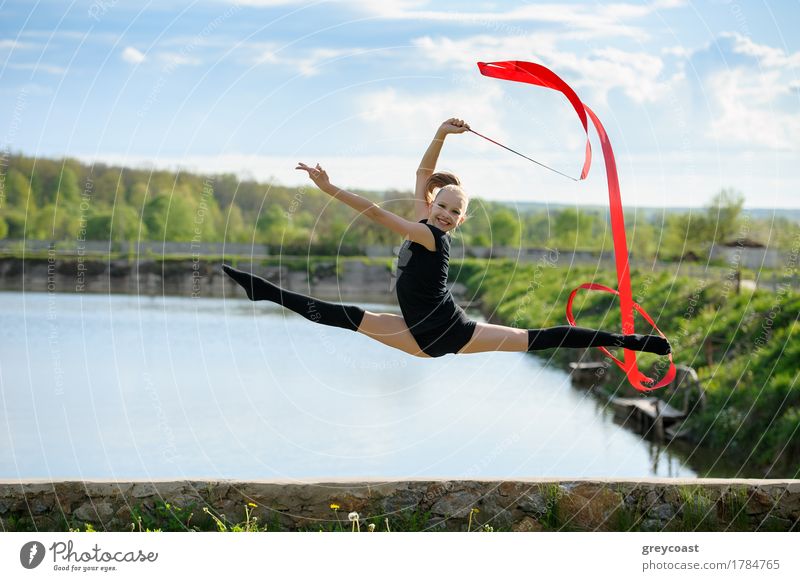 Rhythmische Gymnastin Mädchen Durchführung mit einem roten Band im Freien. Bein-Split in einem Sprung gegen ländliche Natur Hintergrund Glück Körper Sommer