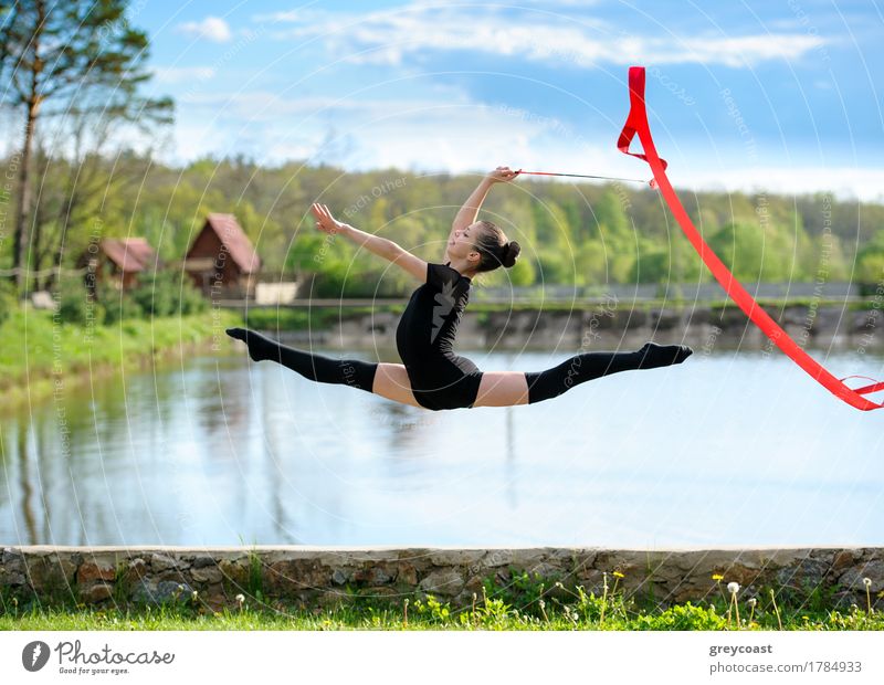 Junge Gymnastin Mädchen tun Übung mit roten Band im Freien. Sie macht Bein-Split in einem Sprung auf dem Hintergrund der Teich und Wald Glück Körper Sommer