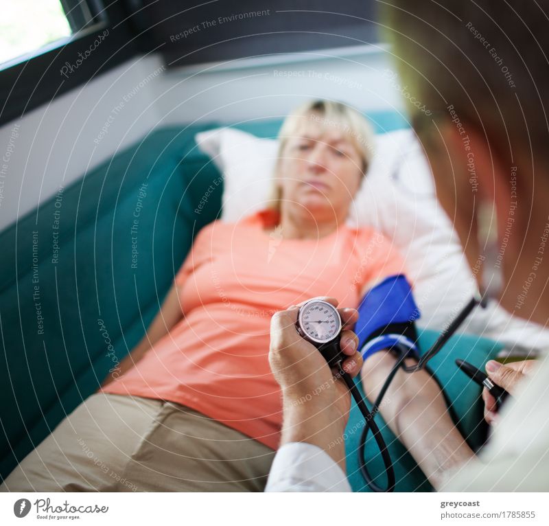 Arzt bei der Blutdruckkontrolle einer Patientin während des Hausbesuchs. Fokus auf Manometer Gesundheitswesen Krankheit Medikament Wohnung Sofa Prüfung & Examen