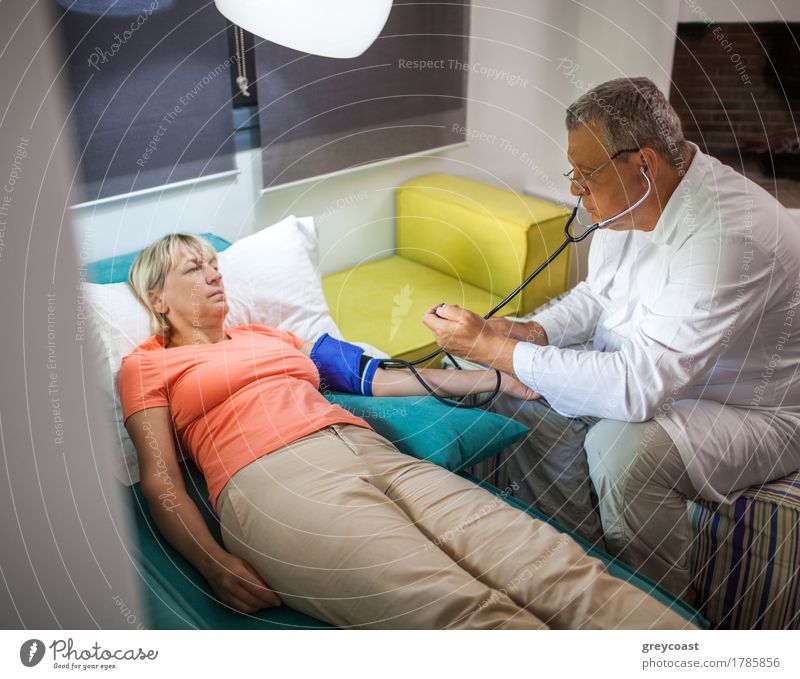 Ältere Frau ist unter ärztlicher Kontrolle zu Hause. Mann prüft Blutdruck mit Tonometer und Stethoskop Gesundheitswesen Krankheit Medikament Wohnung Lampe Sofa