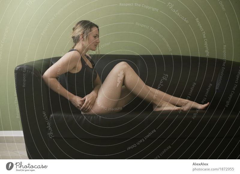 junge, sportliche Frau sitzt in schwarzer Unterwäsche auf einem braunen Sofa vor einer grünen Wand, sie ist sportlich, barfuß, hat lange Beine schön harmonisch