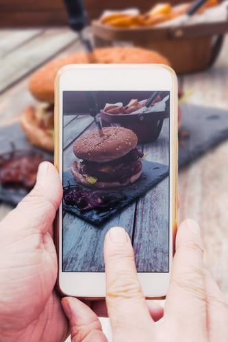 Hand mit Smartphone Handy, Foto von Burger auf Tisch Lebensmittel Fleisch Brot Brötchen Cheeseburger Speck soziale medien teilen Internet Telefon PDA Holz
