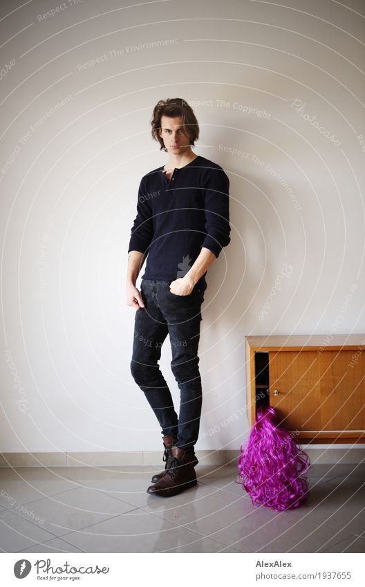junger Mann in einem hellen Raum mit Sideboard und einer lila Perücke darin Lifestyle Möbel Junger Mann Jugendliche 18-30 Jahre Erwachsene rosa Jeanshose