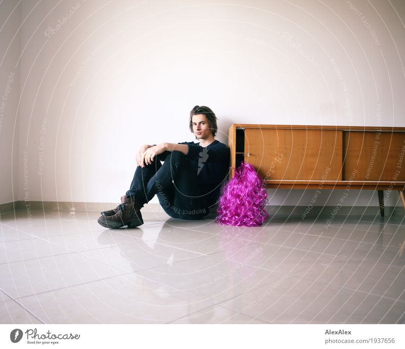 junger Mann sitzt an einem Sideboard in einem hellen Raum neben einer lila Perücke Lifestyle Stil Freude Nachtleben Party Junger Mann Jugendliche 18-30 Jahre