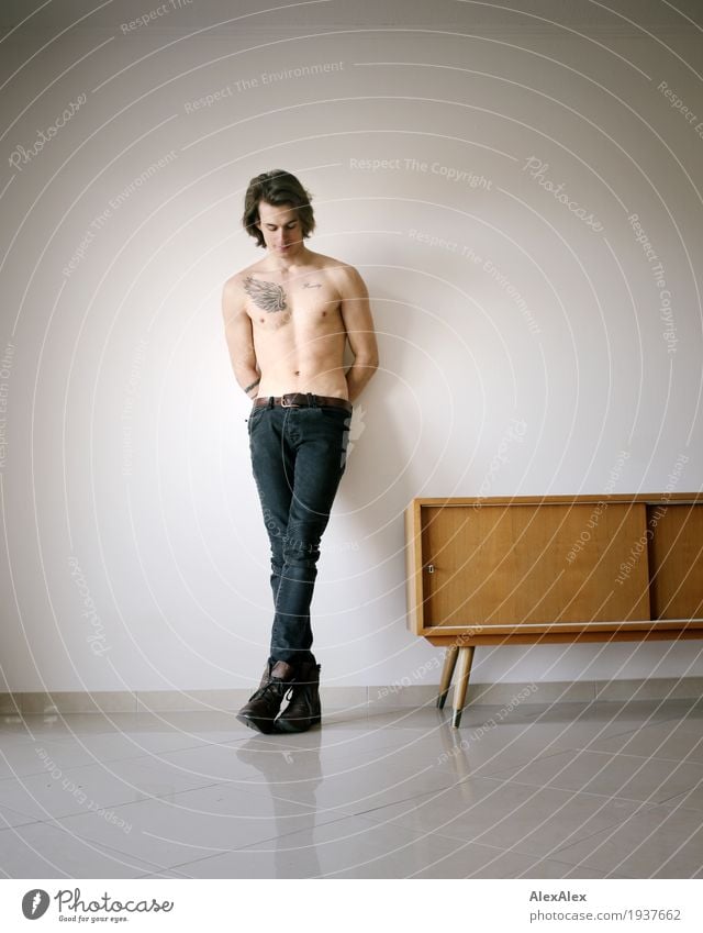 junger Mann mit Tätowierung lehnt mit freiem Oberkörper in einem  hellen Raum neben einem Sideboard Wohnung Möbel Junger Mann Jugendliche nackt 18-30 Jahre