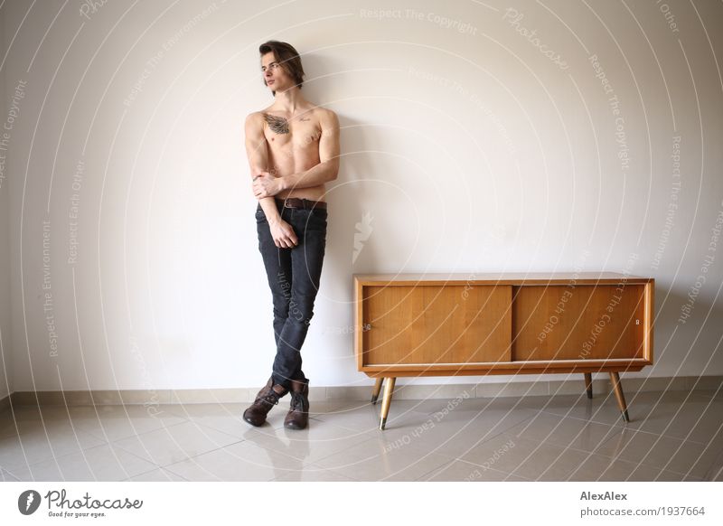 junger Mann mit freiem Oberkörper und Tätowierung lehnt in einem hellen Raum mit Sideboard an der Wand Lifestyle Stil Körper Zufriedenheit Möbel Schrank