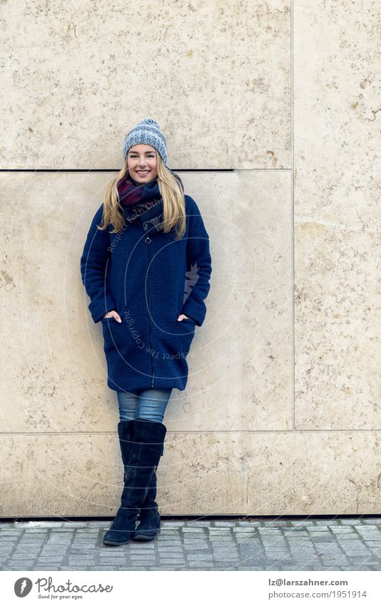 Lächelnde modische junge Frau, die gegen Wand steht Glück schön Erholung Winter Erwachsene Herbst Mode Mantel Schal Stiefel Hut blond stehen dünn trendy lang