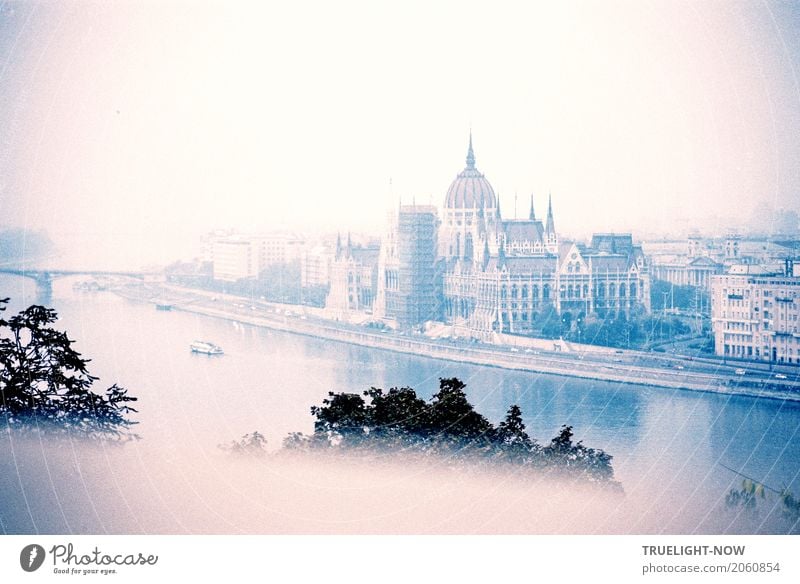 Budapest revisited, Parlament an der Donau, im Morgennebel, vom hochgelegenen Ufer (Buda) gegenüber vereinzelt Schiffe, eine Donaubrücke und im Vordergrund die Spitzen von Baumen