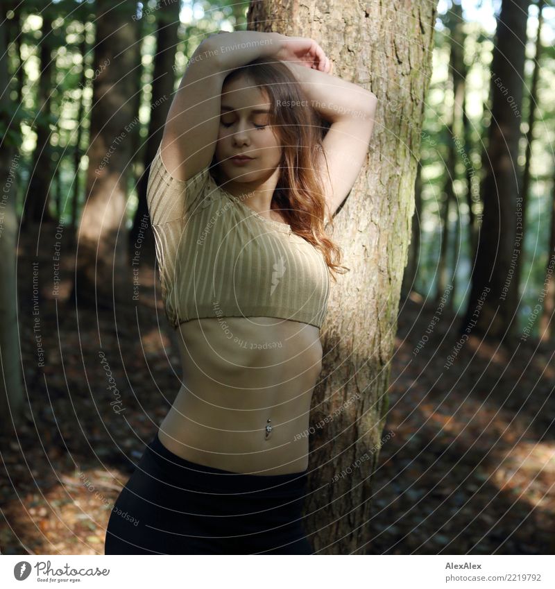 Bauchgefühl - eine junge, sportliche Frau mit einem bauchfreien Top lehnt an einem Baum mit den Armen über dem Kopf verschränkt, geschlossenen Augen und einem Bauchnabelpiercing im muskulösen, flachen Bauch - Licht und Schattenspiel im Wald