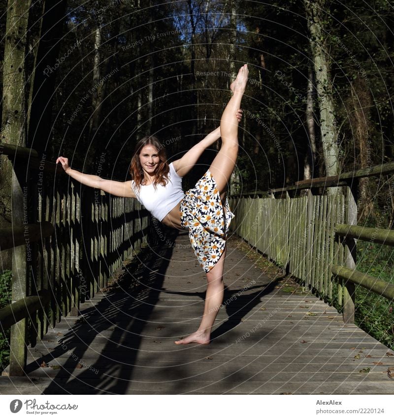 Junge, sehr sportliche Frau mit Sixpack steht barfuß auf einer Holzbrücke im Wald und hält sich ein Bein über den Kopf gestreckt schön Fitness harmonisch Turnen