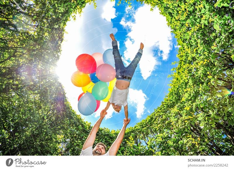 Junge fliegt mit Luftballons. Vater umkreist seinen Sohn hoch über seinem eigenen Kopf. Ringsum hohe grüne Büsche. Klarer blauer Himmel im Hintergrund. Ansicht von unten.