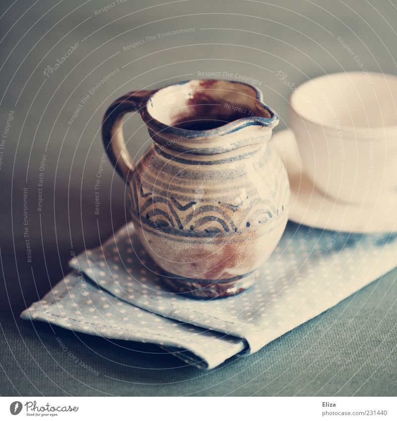 Ein Milchkännchen aus bemalter Keramik neben einer leeren weißen Tasse Milchkanne schön Serviette altehrwürdig blau Geschirr Farbfoto Innenaufnahme Licht