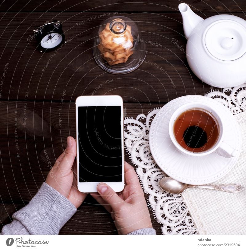 Frauenhände halten ein weißes Smartphone. Frühstück Getränk Tee Teller Becher Lifestyle Tisch Telefon PDA Bildschirm Technik & Technologie Erwachsene Arme Hand