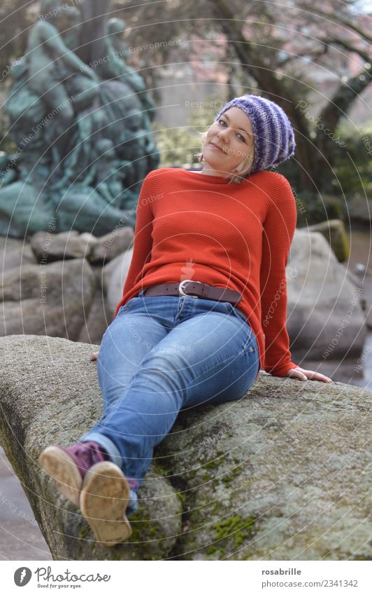junge blonde Frau mit lila Strickmütze sitzt in ihrer Freizeit entspannt und erwartungsvoll in einem Park auf einem Stein und legt den Kopf leicht schräg Freude
