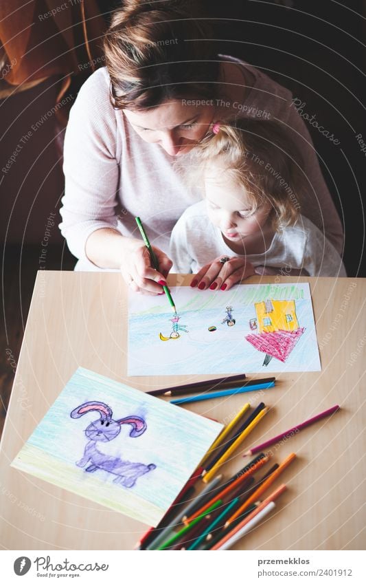 Mutter mit kleiner Tochter malt ein buntes Bild von Haus und spielenden Kindern mit Buntstiften am Tisch sitzend in einem Haus. Aufnahme von oben Lifestyle