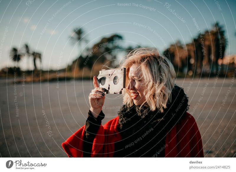 Lächelndes blondes Mädchen, das ein altes kaputtes Videoband in der Hand hält und bei Sonnenuntergang durch die Löcher schaut. Lifestyle Freude Fotokamera
