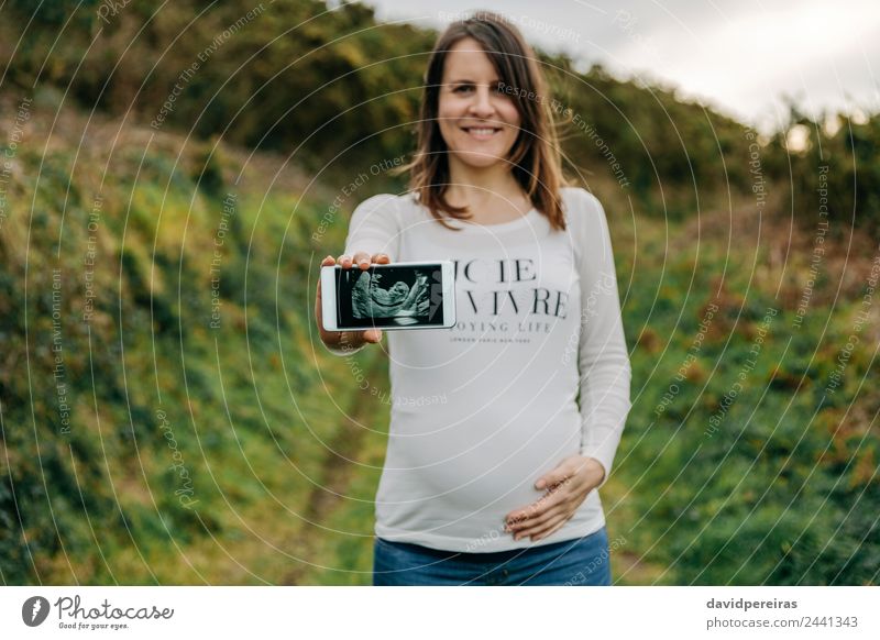 Unbekannte schwangere Frau zeigt Ultraschallbild ihres Babys auf dem Handy Lifestyle Glück schön PDA Mensch Erwachsene Mutter Landschaft Wiese Hemd Lächeln