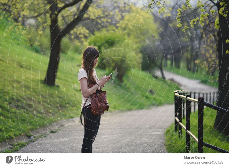 Attraktive junge Frau, die sich draußen in einem Park amüsiert. Kaukasisches Mädchen mit langen braunen Haaren überprüft ihr Handy Lifestyle Glück schön