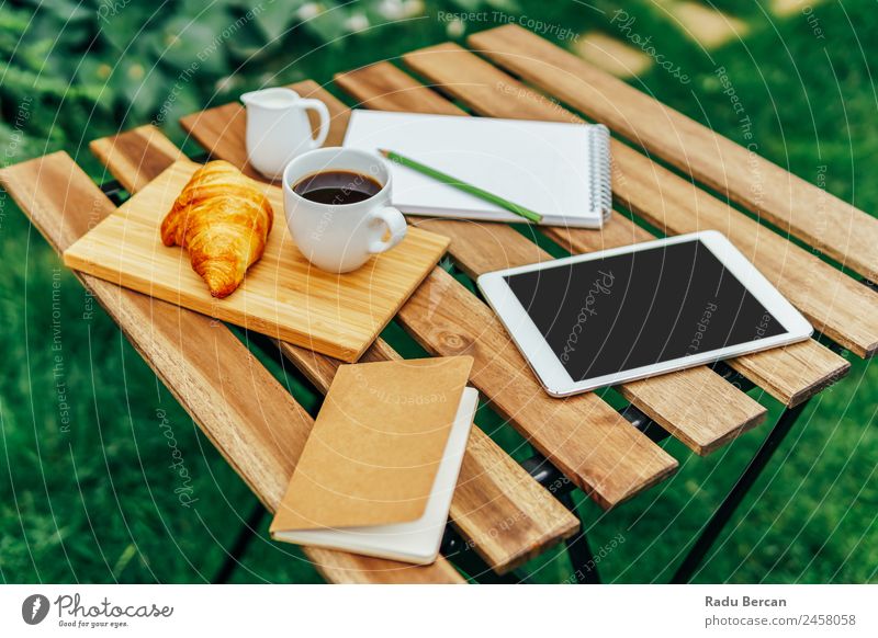 Frühstück am Morgen im Grünen Garten mit französischem Croissant, Kaffeetasse, Orangensaft, Tablette und Notizbuch auf Holztisch Tisch Hintergrundbild Sommer
