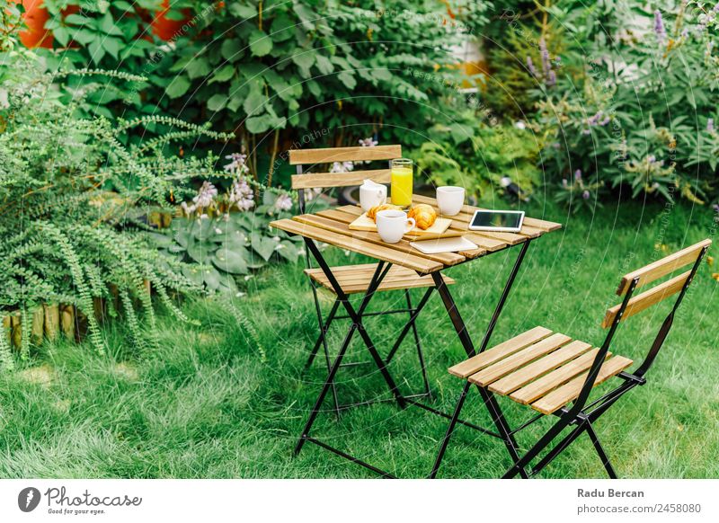 Frühstück am Morgen im Grünen Garten mit französischem Croissant, Kaffeetasse, Orangensaft, Tablette und Notizbuch auf Holztisch Tisch Hintergrundbild Sommer