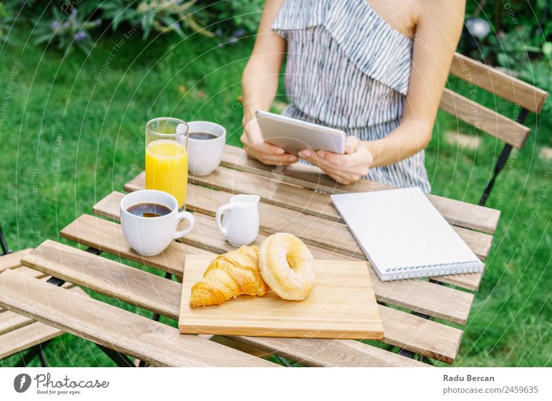 Junge und attraktive Frau beim Frühstück am Morgen im grünen Garten mit französischem Croissant, Donuts, Kaffeetasse, Orangensaft, Tablette und Notizbuch auf Holztisch.